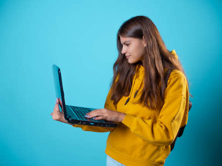 Online training, a young woman uses a laptop. A student with a laptop in her hands, a young brunette in a yellow hoodie on a blue backgroundの写真素材