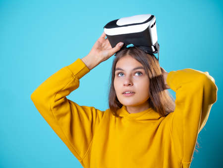 VR gamer, a young woman in a youth outfit uses a virtual reality helmet. A brunette in yellow on a blue background puts a vr gadget on her headの写真素材