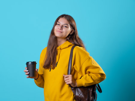 Cute young brunette in jeans and yellow hoodie. A beautiful student holds a takeaway coffee in her hands and carries a backpack on her shoulders, a studio portrait on a blue background.の写真素材
