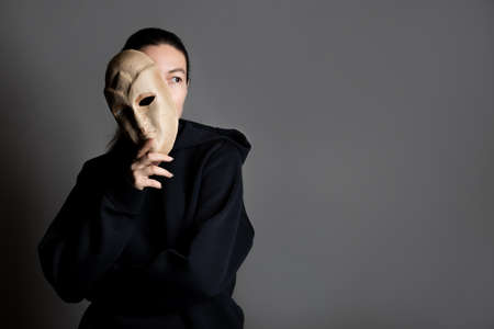 Social masks that people wear in society. Real personality and subpersonalities, social norms and patterns of behavior, psychological concept. young woman hides her face with a mask , portrait on darkの写真素材