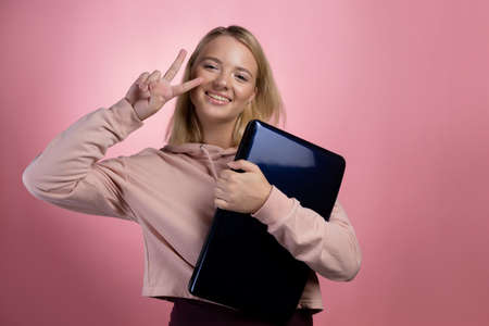 young woman with a laptop, a student or a freelancer. A charming blonde in a pink hoodie on a pink background holds a laptop in her hands and makes a two-finger up gesture.の写真素材