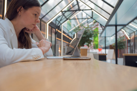 architect or interior designer works remotely. young freelance woman works with an engineering floor plan. brunette in a white jacket with a laptop in public space, drawing of apartment is on screen.の写真素材