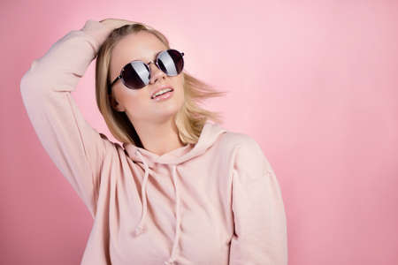beautiful young woman with blonde hair wearing dark sunglasses. In a pink hoodie on a pink background, studio portraitの写真素材