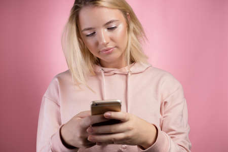 Internet browsing , mobile applications . An attractive young woman in a pink hoodie uses a smartphone. A cute blonde holds a phone and looks at the screen.の写真素材