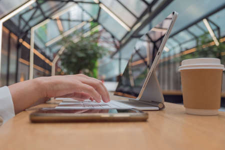 Remote work or freelance, modern opportunities for a mobile lifestyle. Womens hands are typing on keyboard of tablet computer, close-up side view, public space hall on background, low depth of field.の写真素材