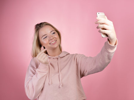 attractive young woman in a pink hoodie uses a smartphone for video communication. Cute blonde shows a gesture of love in the phone screen, the photo is on pink.の写真素材