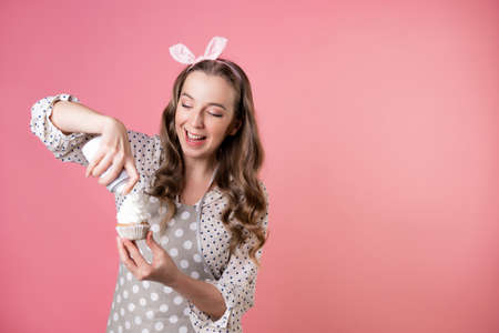 young charming female pastry chef decorates cupcakes with whipped cream. A cook in a polka-dot apron. pastry chef in an apron on a pink background.の写真素材