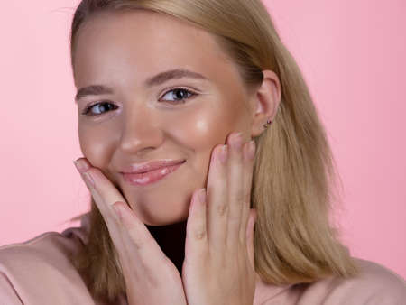 cute young blonde in a pink hoodie smiles happily and touches her face with her hand. portrait on a pink background, close-upの写真素材