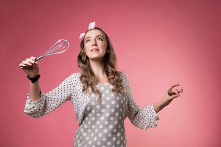 young charming pastry chef woman with a whisk in her hands. Do magic in the kitchen, wave a whisk like a magic wand. A cook in a polka-dot apron. pastry chef in an apron on a pink background.の写真素材