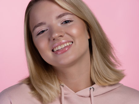 cute young blonde in a pink hoodie is smiling happily. portrait on a pink background, close-upの写真素材