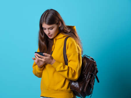 Cute young brunette in jeans and yellow hoodie. smiling student in glasses with a backpack on her shoulders uses the phone to communicate in messengers, a studio portrait on a blue background.の写真素材