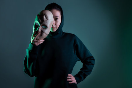 Hiding behind a mask, a young woman in a dark hoodie hides her face with a mask, privacy on the Internet, the concept of information securityの写真素材