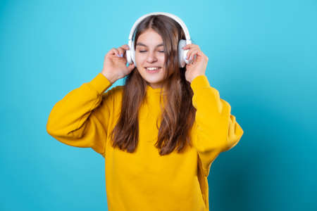 A young smiling student wearing headphones, listening to music. A young brunette in a bright yellow hoodie uses a mobile app to listen to audioの写真素材