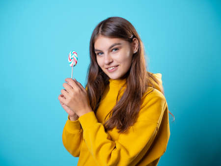 Beautiful young brunette with a lollipop in her hands, sweet candy, portrait in yellow on a blue background, bright colorsの写真素材