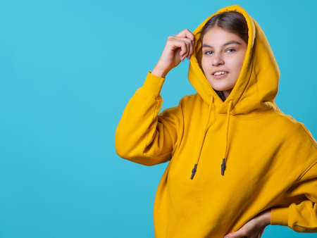 Beautiful young brunette with a hood on her head, youth oversize clothing style, portrait in a yellow hoodie on a blue background, bright colorsの写真素材