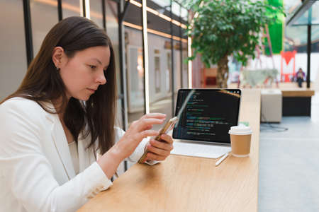 A freelance programmer works remotely, a young woman sits at a table in a coworking or business center and works at a laptop, a business brunette in a white jacketの写真素材