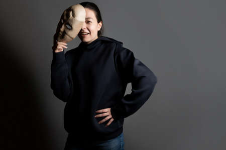 Remove the mask, reveal the identity, express yourself in something, the concept. A young woman in a black hoodie holds a mask in her hands, removing it from her face.の写真素材