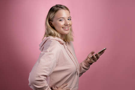 Modern online communication in messengers, correspondence applications. An attractive young woman in a pink hoodie uses a smartphone. A cute blonde holds a phone and looking at youの写真素材