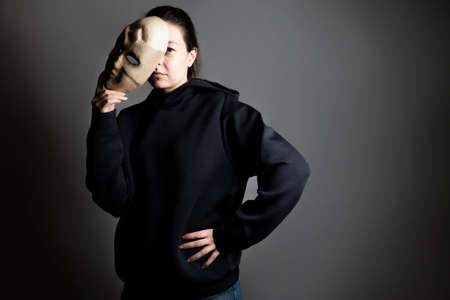 Remove the mask, reveal the identity, express yourself in something, the concept. A young woman in a black hoodie holds a mask in her hands, removing it from her face.の写真素材