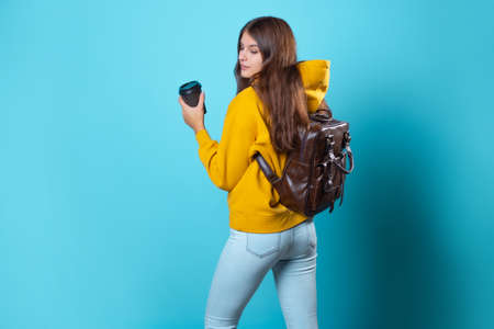 A young student in a yellow hoodie with a cardboard cup of coffee to take away. A girl with a backpack, Portrait on a blue backgroundの写真素材