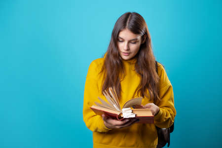 A young student with a backpack looks at a book and reads. Education and training, visiting the library. Young woman in yellow hoodie and jeans on a blue background, copy spaceの写真素材