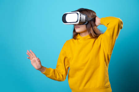 VR gamer, a young woman in a youth outfit uses a virtual reality helmet. Brunette in a yellow hoodie on a blue background with a virtual reality helmet on her head, full immersionの写真素材