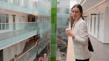 young business woman in a white jacket is standing in a public space with a takeaway coffee in her hand. Confident brunette freelancer, portrait on the background of a business center.の写真素材