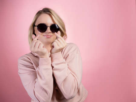 A cheerful young blonde in sunglasses makes a sign of love, shows a heart with her fingers. In a pink hoodie on a pink backgroundの写真素材