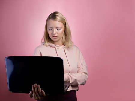 young woman, a student or a freelancer, uses a laptop for work or study. Blonde in a pink hoodie on a pink backgroundの写真素材