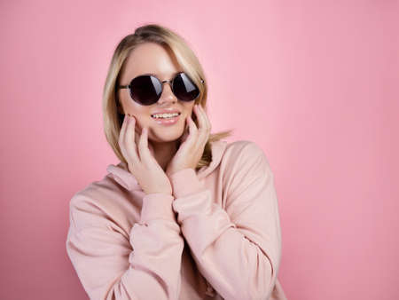 beautiful young woman with blonde hair wearing dark sunglasses. In a pink hoodie on a pink background, studio portraitの写真素材
