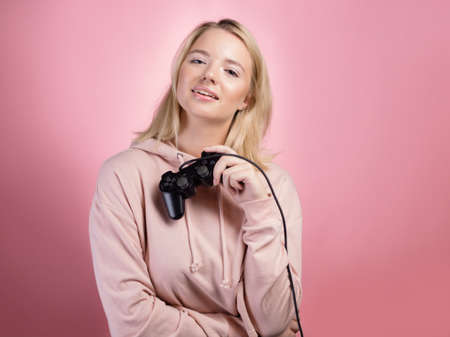 A gamer who is passionate about the process. A cute blonde is holding a gamepad in her hands, looking at the camera. A young woman in a pink hoodie, holding a joystick in her hands ,photo on pinkの写真素材