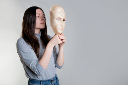 A young woman hides her face behind a mask, the concept of cosmetology and facial skin care, solving skin problems. Photos in light gray tones, gray background.の写真素材