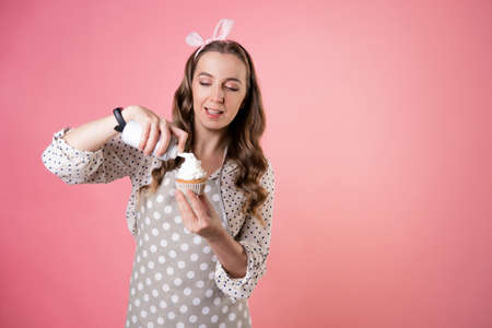 young charming female pastry chef decorates cupcakes with whipped cream. A cook in a polka-dot apron. pastry chef in an apron on a pink background.の写真素材