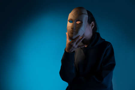 Hiding behind a mask, a young woman in a dark hoodie hides her face with a mask, self-identification problems and impostor syndrome. Portrait in the studio on a dark gray background.の写真素材