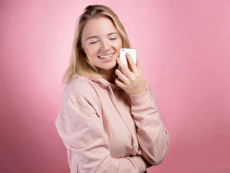 laughing young woman is talking on the phone and smiling . An attractive young woman in a pink hoodie uses a smartphone, a photo on pink.の写真素材