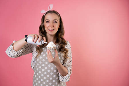 young charming female pastry chef decorates cupcakes with whipped cream. A cook in a polka-dot apron. pastry chef in an apron on a pink background.の写真素材