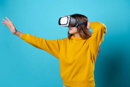VR gamer, a young woman in a youth outfit uses a virtual reality helmet. Brunette in a yellow hoodie on a blue background with a virtual reality helmet on her head, touching something in 3dの写真素材