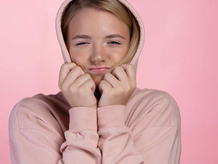 cute young blonde in a pink hoodie puts a hood on her head and squints. portrait on a pink background, close-upの写真素材