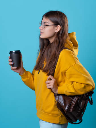 A young student in a yellow hoodie with a cardboard cup of coffee to take away. Young woman with a backpack, Portrait on a blue backgroundの写真素材