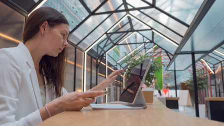 architect or interior designer works remotely. young freelance woman works with an engineering floor plan. brunette in a white jacket with a laptop in public space, drawing of apartment is on screen.の写真素材