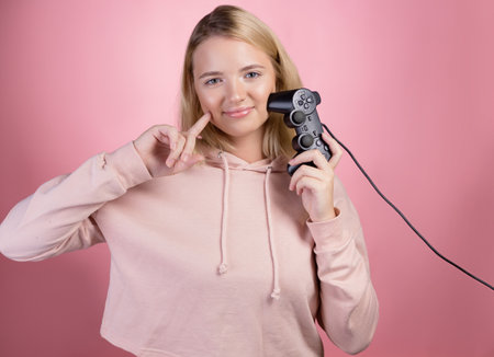 A gamer who is passionate about the process. A cute blonde is holding a gamepad in her hands, looking at the camera. A young woman in a pink hoodie, holding a joystick in her hands ,photo on pinkの写真素材