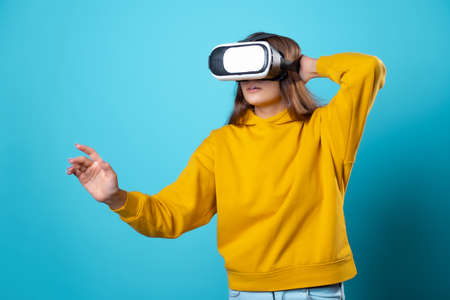 VR gamer, a young woman in a youth outfit uses a virtual reality helmet. Brunette in a yellow hoodie on a blue background with a virtual reality helmet on her head, full immersionの写真素材
