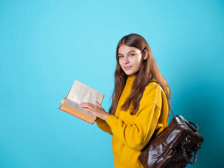 A young student with a backpack looks at a book and reads. Education and training, visiting the library. Young woman in yellow hoodie and jeans on a blue background, copy spaceの写真素材