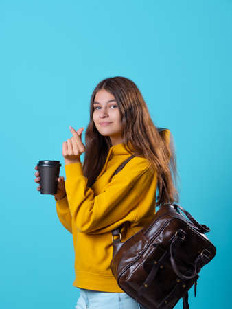 Cute young brunette in jeans and yellow hoodie. smiling student in glasses with a backpack on her shoulders holds a takeaway coffee in her hands, and shows a tin heart, I love.の写真素材
