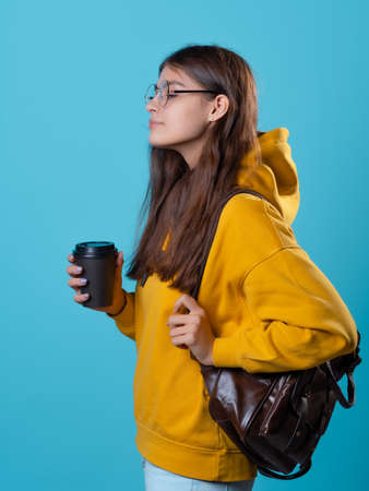 A young student in a yellow hoodie with a cardboard cup of coffee to take away. Young woman with a backpack, Portrait on a blue backgroundの写真素材