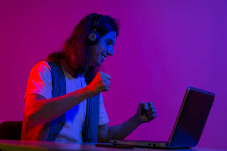 Funny guy gamer playing on a laptop. A young man with headphones is using a computer, playing online games or programming, portrait in neon colorsの写真素材