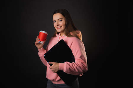 A young cute brunette in a pink hoodie, holding a laptop in her hands and drinking coffee. Student on break, portrait on blackの写真素材