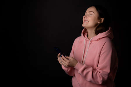Cute young brunette in a pink hoodie uses a smartphone and laughs, communication without borders, concept portrait on a black backgroundの写真素材