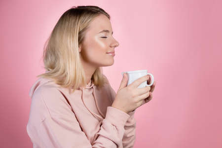 Enjoy your morning coffee, a young positive blonde in pink, holding a white mug in her hands and drinking a drink. Photo on pinkの写真素材