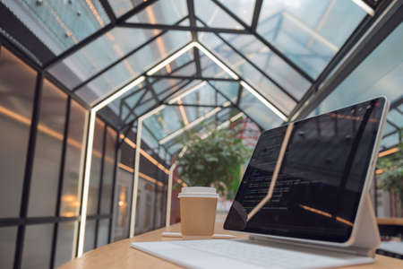 freelancers workplace. A thin mobile laptop on a table in a coworking or other public space. low depth of field. tablet computer with a code on the screen and coffee to take away.の写真素材